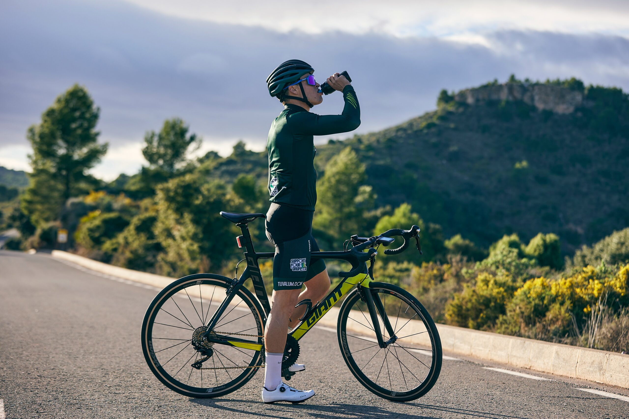 A man drinking energy drink, and standing with cycle.
